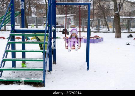 terrain de jeu en hiver.Une petite fille dans une veste violette est assise sur une balançoire et roule autour. Banque D'Images