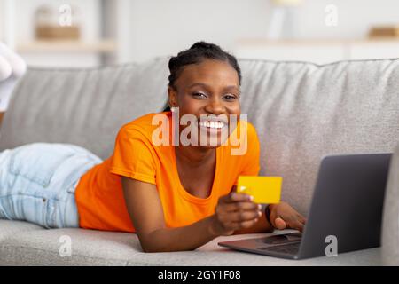 Joyeuse Afro femme magasiner en ligne avec ordinateur portable et carte de crédit, couché sur un canapé à la maison Banque D'Images
