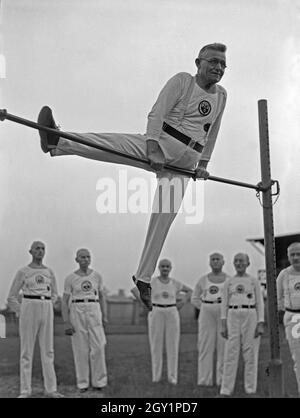 Ein Turner aus der Altherrenriege Turnvereins trägt und eine Übung suis vor Reck, Deutschland 1930er Jahre. Un membre de la haute direction du ministère d'un gymnastic club d'effectuer à un bar sur un terrain de sport, de l'Allemagne des années 1930. Banque D'Images