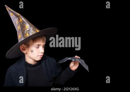 Halloween enfants.Un garçon dans un chapeau de magicien et avec une araignée peinte sur sa joue tenant une batte noire d'Halloween pour effrayer quelqu'un isolé sur un dos noir Banque D'Images