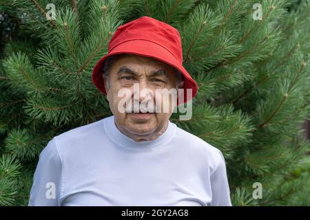 Portrait d'un homme âgé de 70 ans dans un T-shirt blanc et un chapeau rouge qui regarde calmement et attentivement l'appareil photo, dans le jardin de la campagne. Banque D'Images