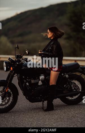 Portrait d'une femme moto assurée en mini jupe, veste en cuir sur une moto de style rétro.Un conducteur attrayant dans des bottes de protection sur route Banque D'Images