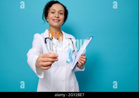 Magnifique médecin de race mixte en robe médicale blanche et ruban de conscience bleu tient un presse-papiers et montre des seringues d'insuline à l'appareil photo, isolé sur bleu Banque D'Images