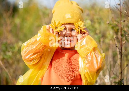 portrait de petite fille drôle en chapeau jaune, chandail orange, imperméable avec de grandes marguerites jaunes couvrant les yeux Banque D'Images