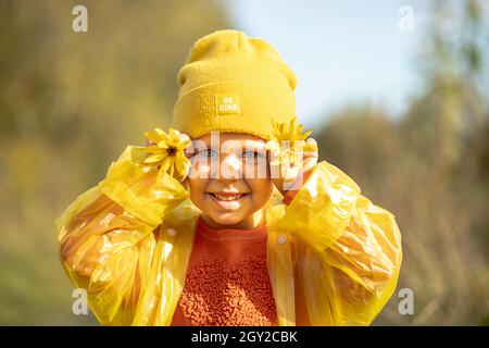 Concept de l'enfance heureuse, portrait de petite fille drôle de 5 ans dans chapeau jaune, chandail orange, imperméable avec grands pâquerettes jaunes couvrant les yeux Banque D'Images