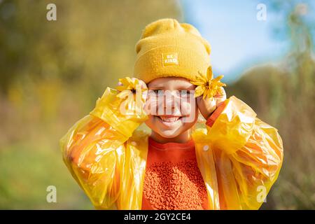 Concept de l'enfance heureuse, portrait de drôle 5 ans fille dans chapeau jaune, chandail orange, imperméable avec grands pâquerettes jaunes couvrant les yeux Banque D'Images