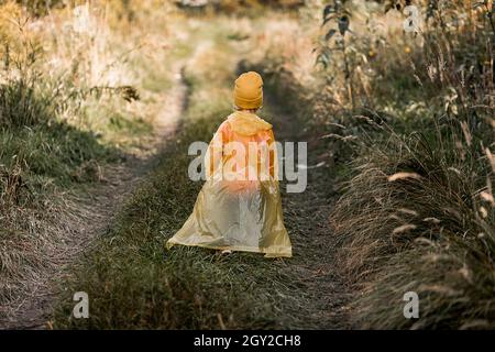 un petit enfant marche le long de la route parmi la grande herbe d'automne en chapeau jaune et imperméable, vue arrière, jour ensoleillé Banque D'Images