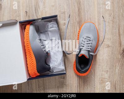 Une sneaker dans la boîte en carton ouverte et l'autre près du sol.Nouvelle paire de sneakers en tissu gris avec semelle rainurée orange.Chaussures en mesh élégantes. Banque D'Images