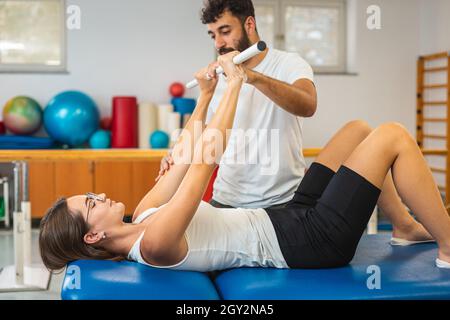 Femme faisant des exercices avec le bar médical pour renforcer ses coudes et ses épaules avec l'aide d'un physiothérapeute masculin Banque D'Images