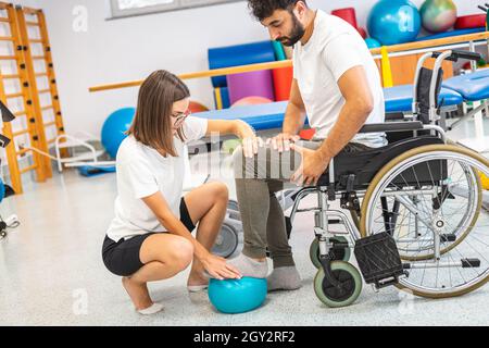 Pkinésithérapeute et homme assis en fauteuil roulant pendant un traitement de réadaptation - appuyer sur un pied et le relâcher sur une petite balle d'exercice Banque D'Images