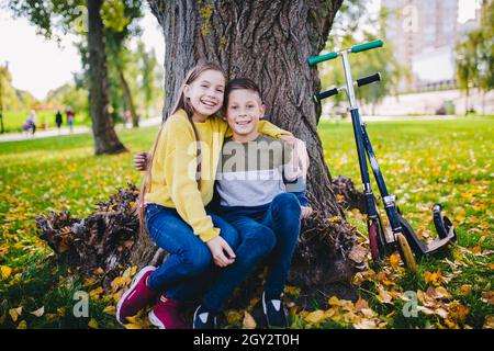 Amis enfants posant heureux assis sous un arbre dans un parc d'automne à côté de kick scooters.Enfants heureux.Transport écologique.Activités de plein air avec lits jumeaux.Bro Banque D'Images