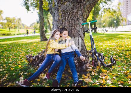 Loisirs actifs et sports de plein air pour les enfants.Bonne enfance.Garçon et fille jumeaux se posant avec joie en s'embrassant tout en étant assis sous un arbre dans un par d'automne Banque D'Images