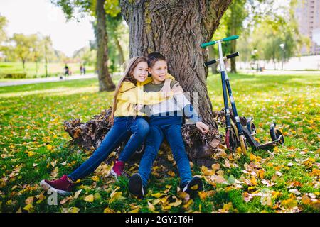 Amis enfants posant heureux assis sous un arbre dans un parc d'automne à côté de kick scooters.Enfants heureux.Transport écologique.Activités de plein air avec lits jumeaux.Bro Banque D'Images