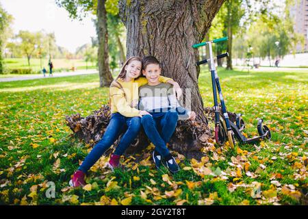 Amis enfants posant heureux assis sous un arbre dans un parc d'automne à côté de kick scooters.Enfants heureux.Transport écologique.Activités de plein air avec lits jumeaux.Bro Banque D'Images