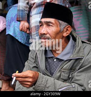 Un vieil homme portant un chapeau de peci noir musulman tout en fumant une cigarette dans un marché à Bukittinggi, Sumatra Ouest, Indonésie. Banque D'Images