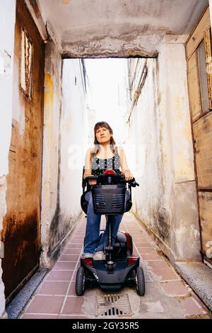Jeune femme sur un fauteuil roulant de scooter passant par un couloir rustique et un inconnu Banque D'Images