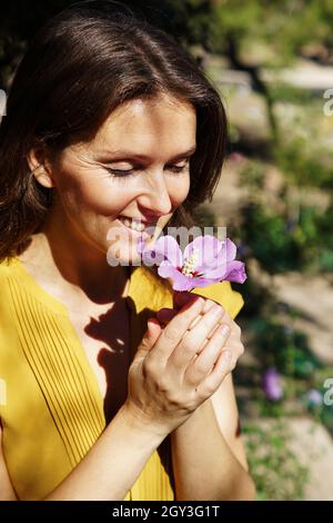 Jeune femme espagnole souriante sentant une fleur d'hibiscus violet frais dans un parc Banque D'Images