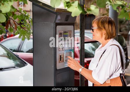 Femme d'âge moyen souriante utilisant une machine à garer pour payer le parking dans la rue d'été de la ville Banque D'Images