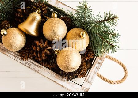 Jouets de Noël, boules d'or et cônes forestiers dans une boîte en bois blanc avec poignées en corde sur fond en bois blanc. Vue du dessus. Angle étroit Banque D'Images