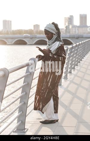 Femme musulmane africaine en vêtements traditionnels lisant un message sur un téléphone portable tout en se tenant debout sur le pont à l'extérieur Banque D'Images