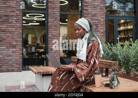Femme d'affaires musulmane africaine assise sur le banc avec ordinateur portable et workikng en ligne à l'extérieur Banque D'Images
