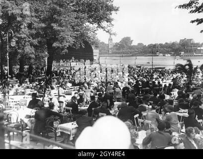 Lors d'une belle journée à l'été 1931, le restaurant Spreegarten du parc Treptower de Berlin est très fréquenté.Un orchestre donne un concert sur la scène.En arrière-plan, de l'autre côté de la Spree, on peut voir l'église du village de Stralau (au centre de l'image).[traduction automatique] Banque D'Images