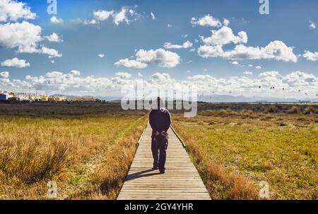 Homme marchant le long d'une promenade en bois à travers des prairies vers des surfeurs de cerf-volant éloignés dans un ciel ensoleillé et nuageux avec des bâtiments blanchis à la chaux sur le côté dans un Banque D'Images