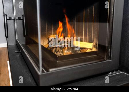Bois brûlé dans une cheminée moderne avec une chambre de combustion fermée dans le salon, peint en noir, avec une vitre d'angle. Banque D'Images