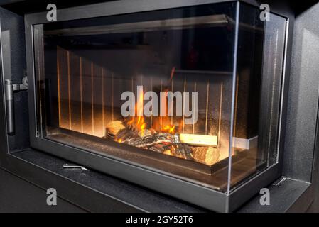 Bois brûlé dans une cheminée moderne avec une chambre de combustion fermée dans le salon, peint en noir, avec une vitre d'angle. Banque D'Images