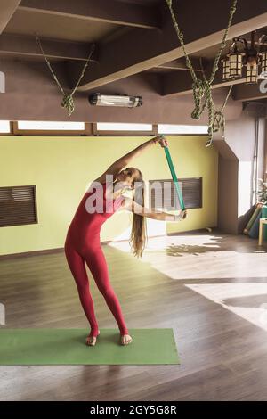Une femme pratiquant la thérapie de yoga, effectue des coudes latéraux avec une ceinture, se tient sur un tapis dans une combinaison rouge de sport dans le studio Banque D'Images