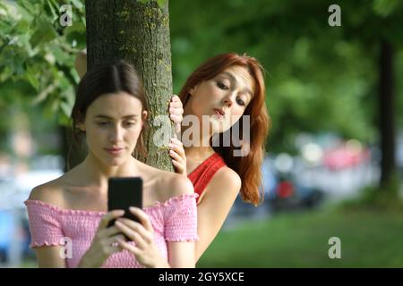 Une femme distraite en utilisant un smartphone et est espionné par son amie curieuse dans un parc Banque D'Images