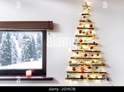 Sapin de Noël en bois suspendu au mur, paysage enneigé d'hiver à l'extérieur de la fenêtre. Banque D'Images