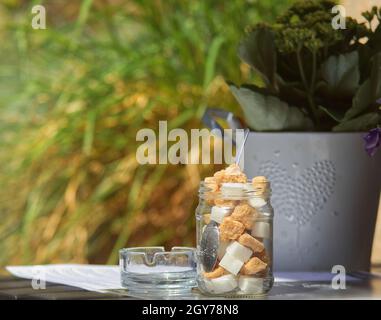 Un pot de sucre en cube, de sucre en cube brun et blanc dans un pot sur une table basse dans la rue, avec un petit cendrier à côté.Arrière-plan flou. Banque D'Images