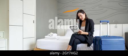 Femme d'affaires asiatique en costume noir assise avec un ordinateur tablette dans la main. Ses bagages ont été placés à côté du lit. Atmosphère de travail à l'extérieur pendant le Banque D'Images