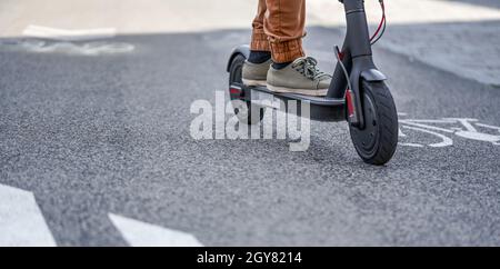 Jeune homme dans un pantalon décontracté, portant un détail de scooter électrique sur ses pieds et roue au-dessus de la route d'asphalte. Banque D'Images