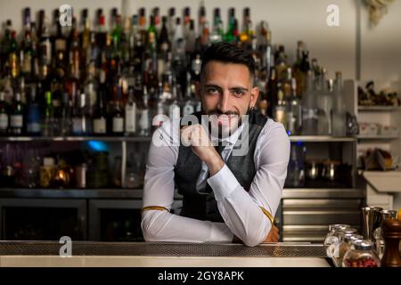 Beau jeune barbu hispanique homme barbu dans des vêtements élégants souriant amical et regardant la caméra tout en se tenant derrière le comptoir de bar avec alcoho Banque D'Images