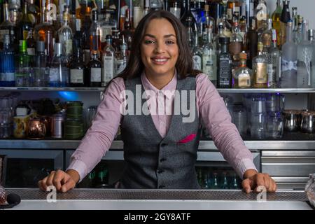 Bonne jeune femme hispanique en chemise et gilet souriant et regardant la caméra tout en se tenant derrière le comptoir contre la tablette avec de l'alcool pendant le travail Banque D'Images