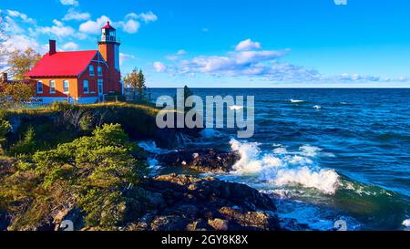 Phare d'Eagle Harbor avec vagues de lac écrasant et arbres verts avec un ciel bleu magnifique et des nuages blancs et violets Banque D'Images