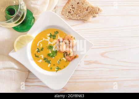 Soupe de crème de légumes de fête à base de courge rouge kuri avec crevettes et garniture de persil dans une élégante assiette blanche, pain et vin sur une table en bois clair, Banque D'Images