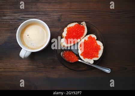 Sandwiches avec caviar rouge, tasse à café, assiette et cuillère sur fond de bois. Pose à plat. Banque D'Images