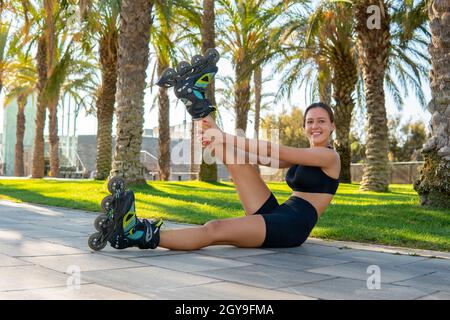 Vue latérale d'une athlète féminine heureuse en rollers levant et étirant la jambe tout en étant assis sur la chaussée pendant l'entraînement dans le parc tropical. Banque D'Images