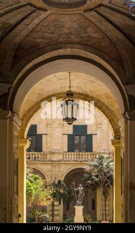 Entrée élégante en forme d'arche dans la cour de la Valetta Malte avec statue, palmiers, plafond orné, lampe de suspension et personne. Banque D'Images