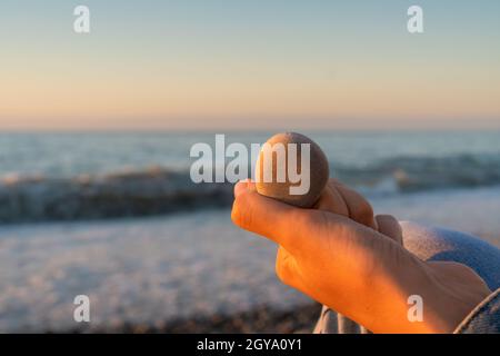 La main de la femme tient une pierre de mer sur le fond du ciel, la mer avec des vagues, une plage de galets au coucher du soleil de près Banque D'Images