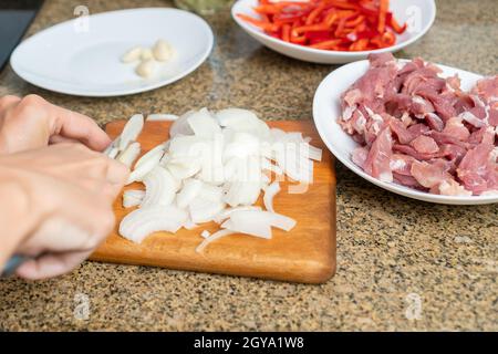 Gros plan des mains des femmes coupant des oignons blancs sur une planche à découper dans la cuisine, à côté des assiettes avec des morceaux de viande de porc, poivron rouge doux Banque D'Images