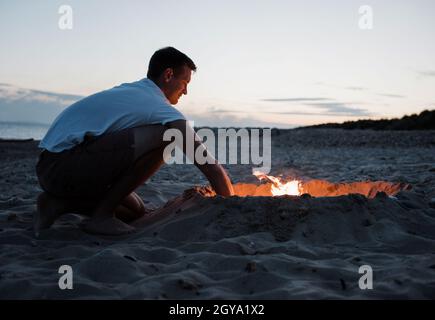 homme heureux de commencer un feu de joie à la plage au coucher du soleil Banque D'Images