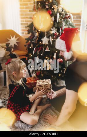 Un jeune père en chapeau de père Noël rouge donne un cadeau de Noël à sa fille souriante près d'un arbre de Noël décoré. Fille vêtue de Noël rouge-noir festif Banque D'Images