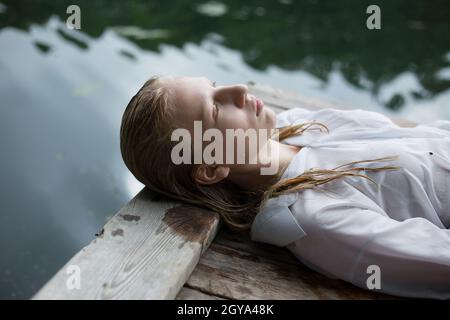 Une adolescente blonde en chemise blanche aux cheveux mouillés est couchée au bord du lac Banque D'Images