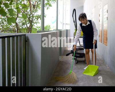 Une jeune femme de ménage nettoie le sol avec un balai et une pelle à poussière. Chariot de ménage dans le couloir devant la chambre. Le panier à linge et le système d'aspiration cl Banque D'Images