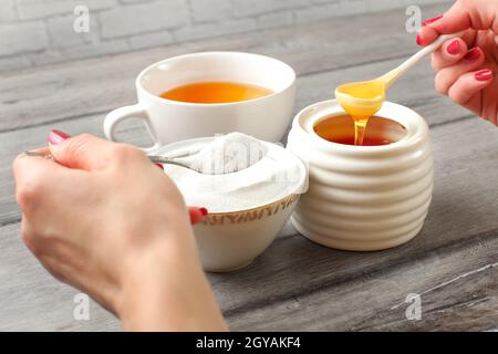 Jeune femme mains tenant cuillérée de sucre, et le miel de décider quoi mettre dans le thé. L'édulcorant naturel raffiné concept. Banque D'Images