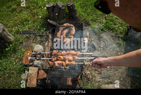 Homme préparant du shashlik ou shish kebab sur le charbon de bois. Faites griller de la viande et des saucisses ou de la bratwurst sur une grille à grillades dans l'arrière-cour. Viande grillée sur brochettes métalliques Banque D'Images